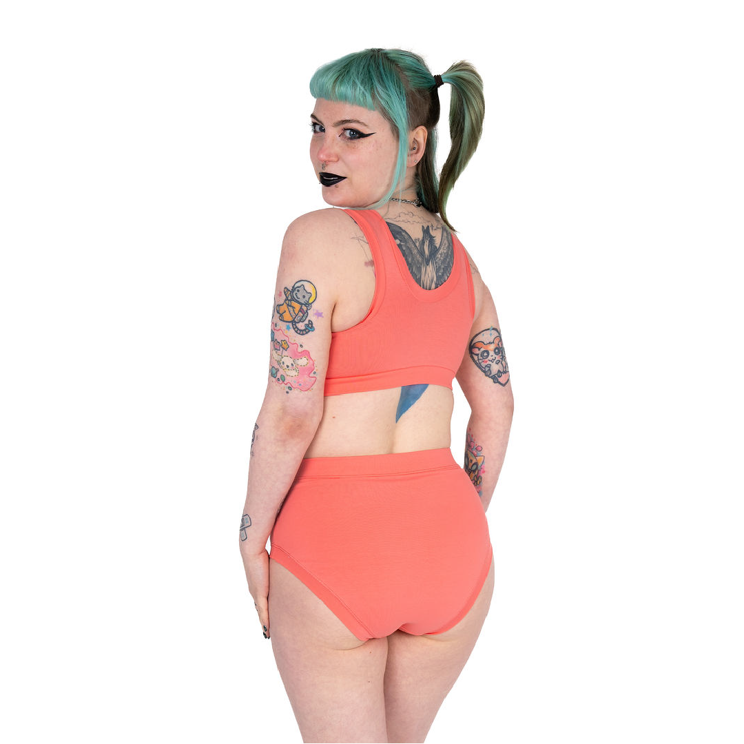Facing away from camera and looking over their shoulder, Faeryn wearing a coral bra and high rise briefs