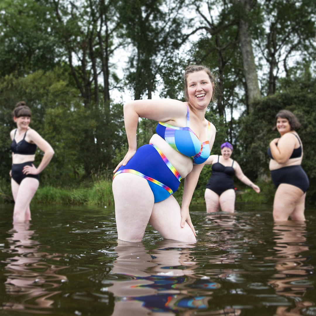 Katy is posing in a blue rainbow bikini with friends in black swimwear in the background laughing